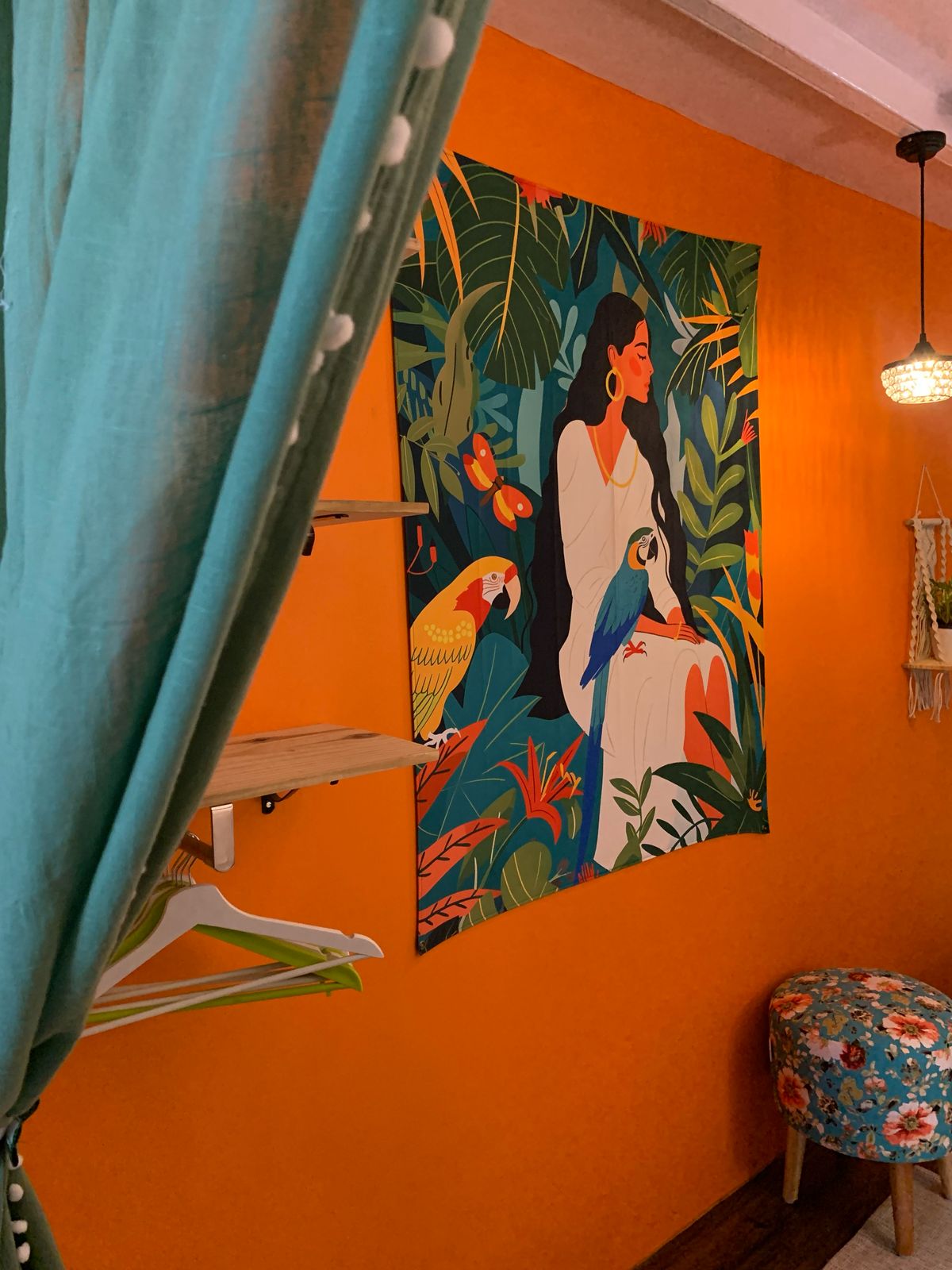 A wall art tapestry featuring a stylized woman figure with a parrot, surrounded by tropical leaves, displayed on an orange wall with a blue curtain to the side.